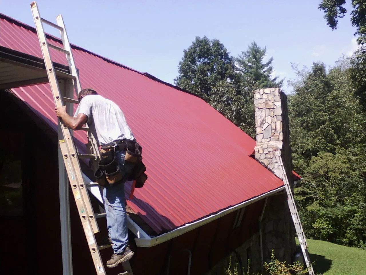 Roofer on a ladder beside a red metal roof and stone chimney — Soffit & Fascia Repair by Roofing Brothers in West Springfield